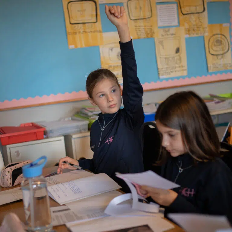 two school girls at a desk, one raising her arm